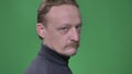 Portrait of middle-aged serious man with beard turns to camera and watches with sneer on green background.