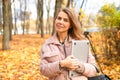 Portrait of middle-aged woman standing near trees on yellow fallen maple leaves in park forest in autumn holding laptop. Royalty Free Stock Photo