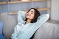 Portrait of millennial Indian lady resting on sofa at home, closing her eyes, putting hands behind head