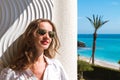 Portrait of a modern long hair woman with sunglasses smiling and standing indoor on the balcony with seascape view.