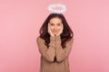 Portrait of modest young pretty woman with halo above head holding hands on face and looking at camera