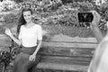 Happy beautiful woman smiling while sitting on wooden bench with Indian man taking picture with mobile phone at peaceful Royalty Free Stock Photo