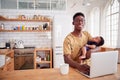 Portrait Of Multi-Tasking Father Holding Sleeping Baby Son And Working On Laptop Computer In Kitchen Royalty Free Stock Photo