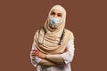 Portrait of Muslim doctor woman wearing white robe and protective mask, arms crossed, standing near brown background. The concept Royalty Free Stock Photo