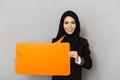 Portrait of muslim smiling woman 20s in black traditional clothing looking at camera while holding orange empty copyspace bubble Royalty Free Stock Photo