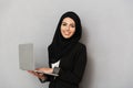 Portrait of muslim smiling woman 20s in black traditional clothing looking at camera while holding laptop, isolated over gray Royalty Free Stock Photo