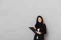 Portrait of muslim smiling woman 20s in black traditional clothing looking at camera while holding clipboard with Royalty Free Stock Photo