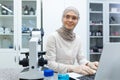 Portrait of Muslim young woman in hijab, chemist scientist working in laboratory at laptop and writing information and