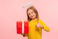 Portrait of nice kind little ginger girl with freckles and angelic halo over head holding gift box and showing thumb up