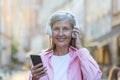 Portrait of an older beautiful gray-haired woman wearing headphones listening to music on the phone. Standing on a city Royalty Free Stock Photo