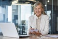 Portrait of an older gray-haired happy business woman working in the office. He sits with a laptop in a suit, holds a Royalty Free Stock Photo