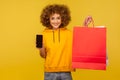 Portrait of optimistic curly-haired hipster girl in hoodie holding cellphone and shopping bags with blank area
