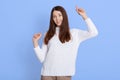 Portrait of optimistic dark haired young woman poses with hands up, dances carefree, wears white jumper, looks at camera, isolated