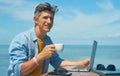 Portrait optimistic smiling man freelancer working outdoors on beach by blue sea, drinking coffee Royalty Free Stock Photo