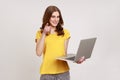 Portrait of optimistic young woman in yellow T-shirt holding laptop, pointing finger to camera and Royalty Free Stock Photo