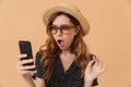 Portrait of outraged confused woman wearing straw hat and sunglasses holding and looking at mobile phone, isolated over beige Royalty Free Stock Photo