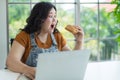 Portrait of overweight asian woman sitting and holding a hamburger open her mouth and hesitate of eating while using a laptop