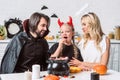 portrait of parents and daughter in halloween costumes at table with treats in black pot in kitchen Royalty Free Stock Photo