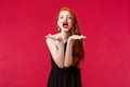 Portrait of passionate and sassy, seductive young beautiful woman with red hair in black dress, sending air kiss to Royalty Free Stock Photo