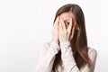 Portrait of peeping pretty young woman in light clothes hiding, covering face eith hands isolated on white wall