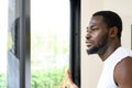 Portrait of pensive African American man standing, touching, and looking outside the window at home Royalty Free Stock Photo