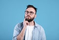 Portrait of pensive bearded man in glasses touching his chin, deep in thought over blue studio background Royalty Free Stock Photo