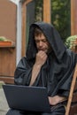 Portrait of a pensive elderly monk 45-50 years old in a black cassock sitting at a laptop against the background of the city. Conc