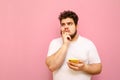 Portrait of pensive fat man standing with smartphone in hands on pink background, looking away at copy space and thinking. Curly