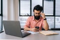 Portrait of pensive Indian businessman in glasses using mobile phone texting online message sitting at office desk with Royalty Free Stock Photo