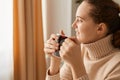 Portrait of pensive positive woman with ponytail hairstyle wearing beige casual style sweater standing near window with cup of