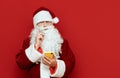 Portrait of pensive santa claus on red background holding smartphone and looking into camera. Funny Santa uses smartphone isolated Royalty Free Stock Photo