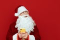 Portrait of pensive santa claus standing on red background with smartphone in hand and looking away at blank copyspace. Royalty Free Stock Photo