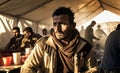 Portrait of pensive and serious young man sitting inside of a temporal tent homes in a refugee camp with people eating and cooking