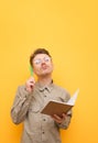 Portrait of pensive student on yellow background with exercise book and pen in hand on yellow background, looking up and thinking Royalty Free Stock Photo