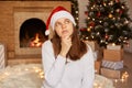 Portrait of pensive woman wearing white sweater and santa claus hat, sitting near fireplace and xmas tree in festive living room, Royalty Free Stock Photo