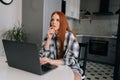 Portrait of pensive young woman sitting at table with laptop computer, pensive looking away in kitchen interior Royalty Free Stock Photo