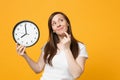 Portrait of pensive young woman in white casual clothes put hand prop up on chin, holding round clock isolated on yellow Royalty Free Stock Photo