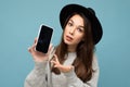 Portrait photo shot of beautiful young woman wearing black hat and grey sweater holding phone showing smartphone Royalty Free Stock Photo