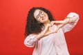 Portrait of a smiling young curly woman showing heart gesture with two hands and looking at camera isolated over red background Royalty Free Stock Photo