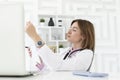 Portrait photo of young and beautiful smiling female doctor in white medical gown sitting and working on laptop Royalty Free Stock Photo