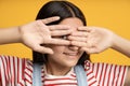 Portrait playful smiling teen girl hiding looking through hand one eye on yellow background.