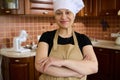 Pleasant housewife in chef's hat and apron, standing with arms crossed in the home kitchen, smiling, looking at
