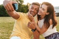 Portrait of pleased adult couple showing thumb up and taking selfie photo on cellphone while sitting on grass in park