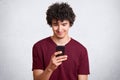 Portrait of pleased attractive man with curly hair and little beard holding smartphone and staring at its scren with intrigued