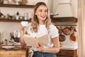 Portrait of pleased blond woman reading book and drinking tea while standing in stylish wooden kitchen at home Royalty Free Stock Photo