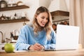 Portrait of pleased blond woman 20s wearing casual sweatshirt working on laptop and writing down notes at home Royalty Free Stock Photo