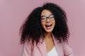 Portrait of pleased young curly haired woman feels overemotive, keeps mouth widely opened, wears optical glasses and formal suit,