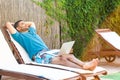 Portrait of pleasure bearded young adult freelancer man in blue t-shirt and shorts lying on cozy sunbed with laptop and