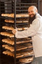 Portrait of pleasure young adult baker with long beard in white uniform standing near of the shelves full with fresh baked cookies Royalty Free Stock Photo