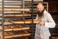 Portrait of pleasure young adult baker with long beard in white uniform standing near of the shelves full with freshly baked bread Royalty Free Stock Photo